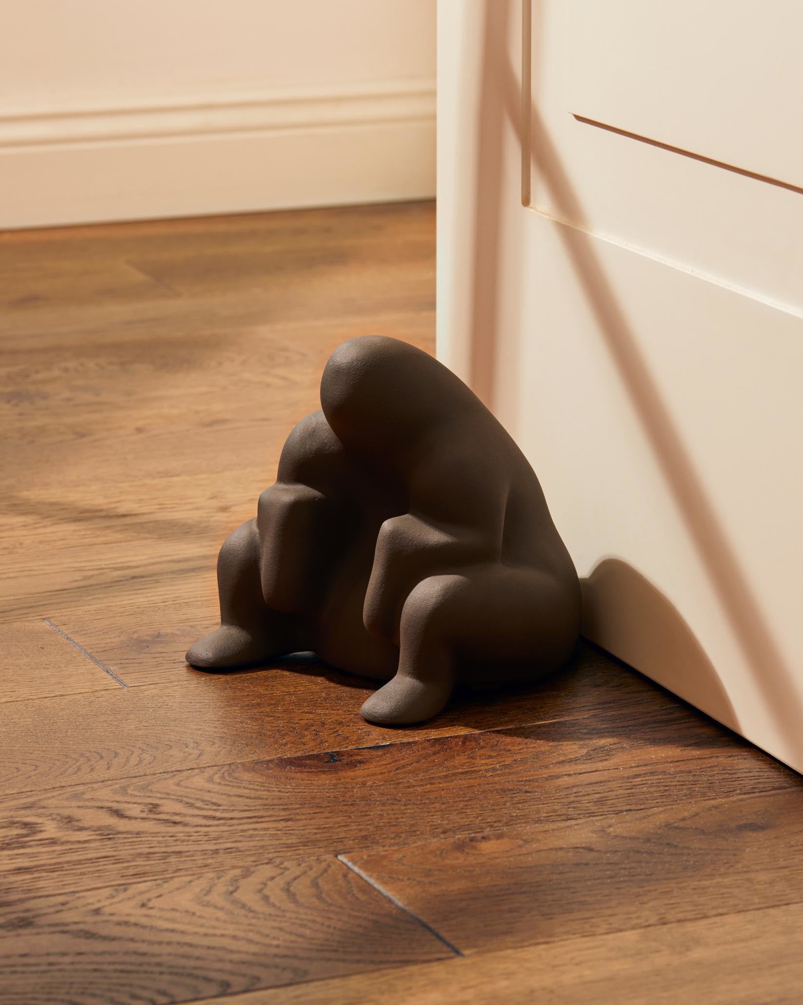 Matte dark brown Dédé doorstop by Alessi, an abstract crouching figure propping open a white door on hardwood.