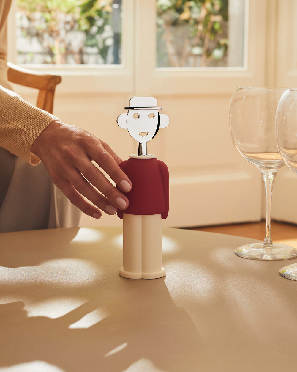 Figural corkscrew with chrome head, red shirt, and cream pants by Alessi, a woman's hand reaching for it on a table with wine glasses.