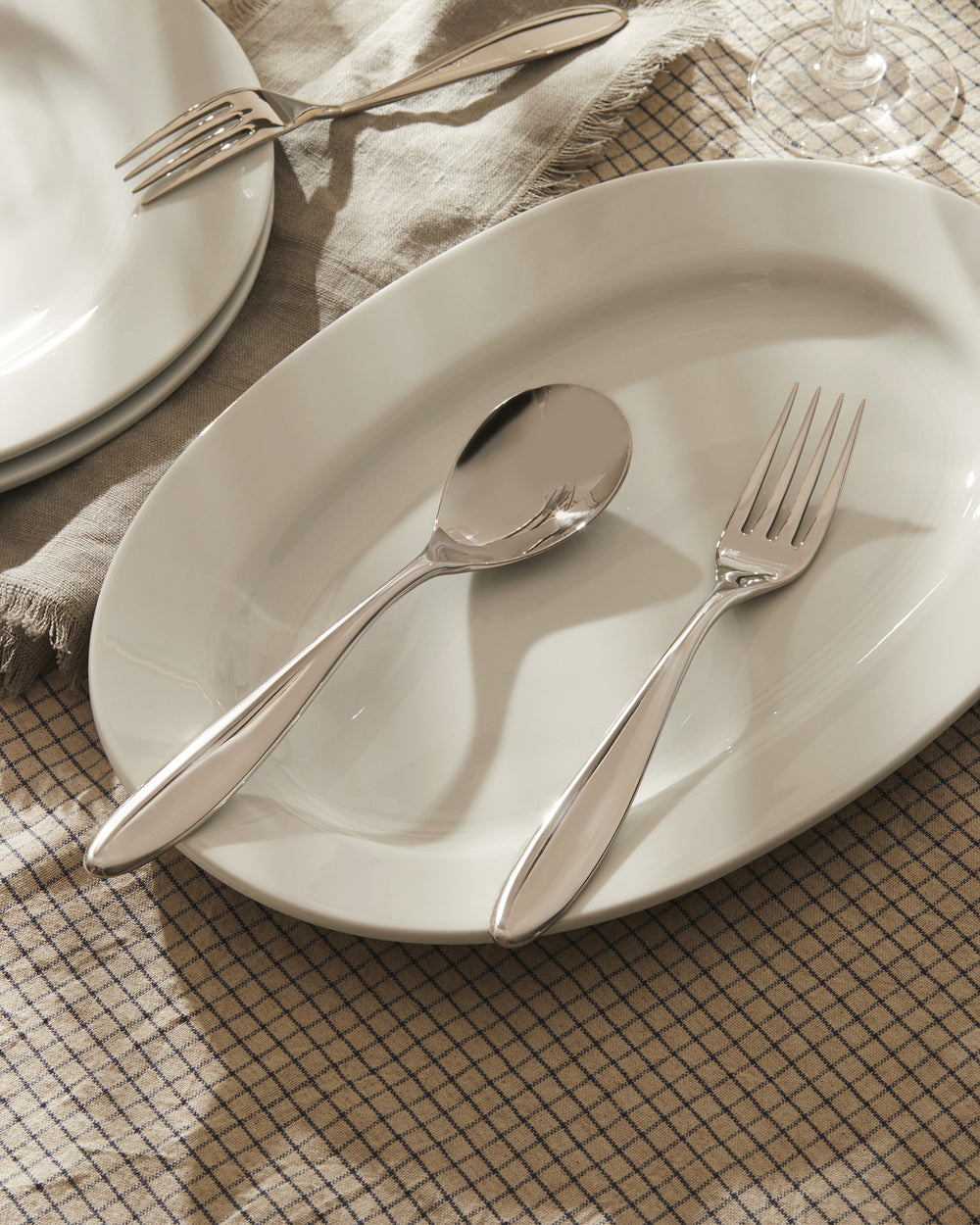 Silver serving fork by Alessi, with a matching spoon on a white oval platter and grid-patterned tablecloth.
