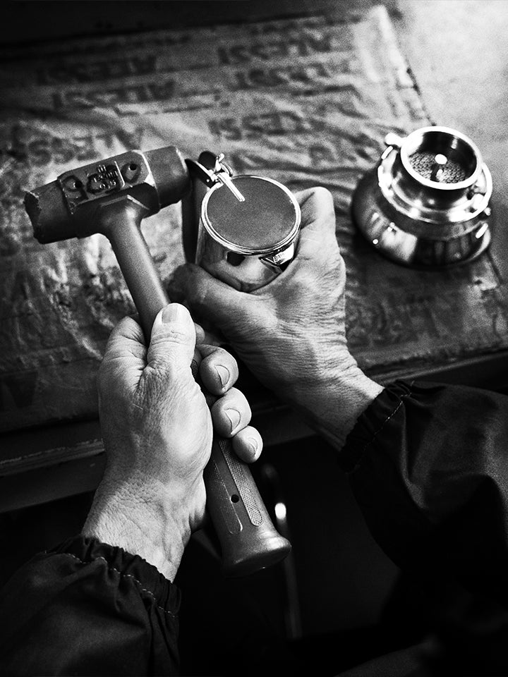 Hands holding a hammer and metal container over newspaper, with a lantern nearby in black and white.