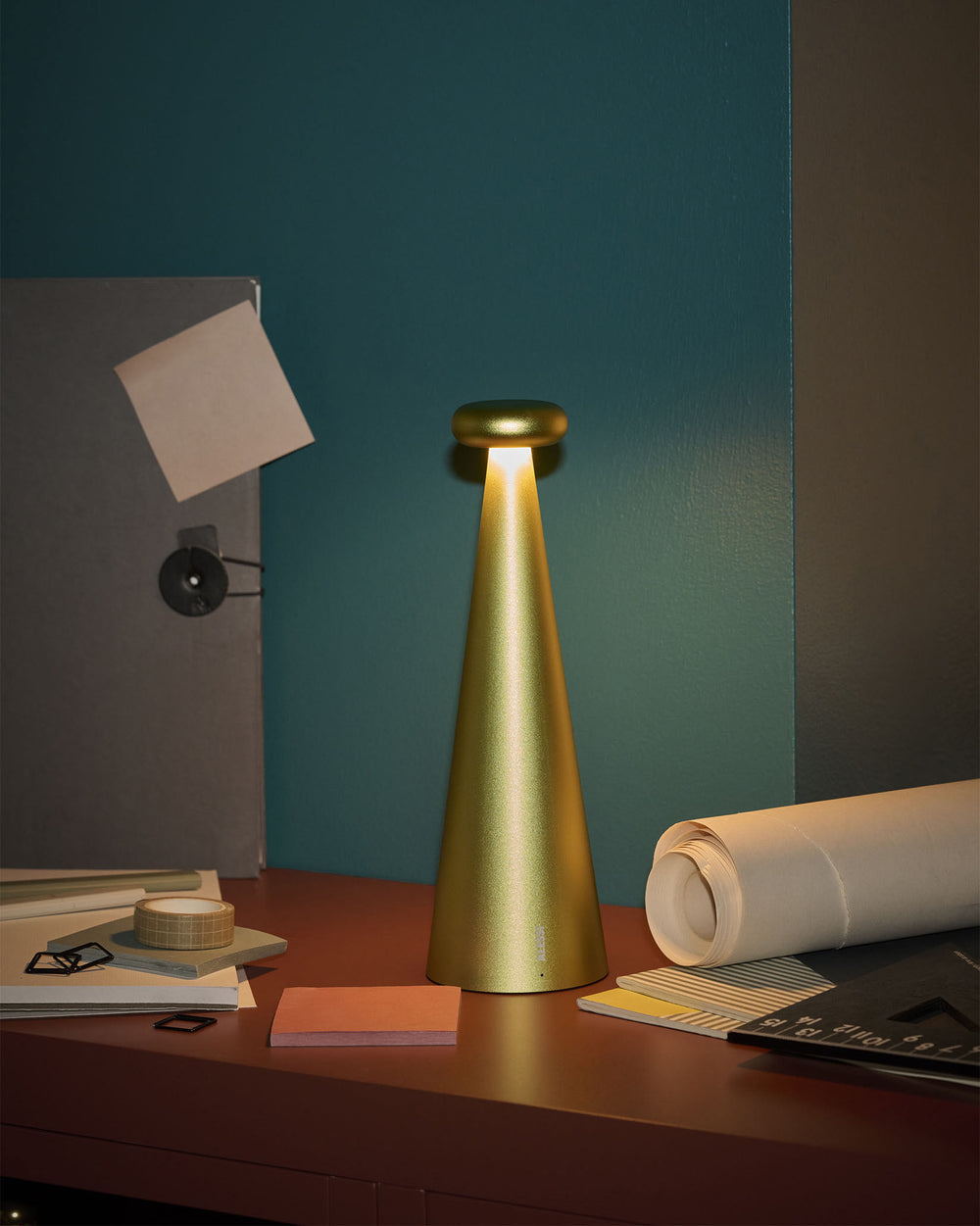Gold brushed conical lamp by Alessi with a rounded top, emitting warm light, on a desk.