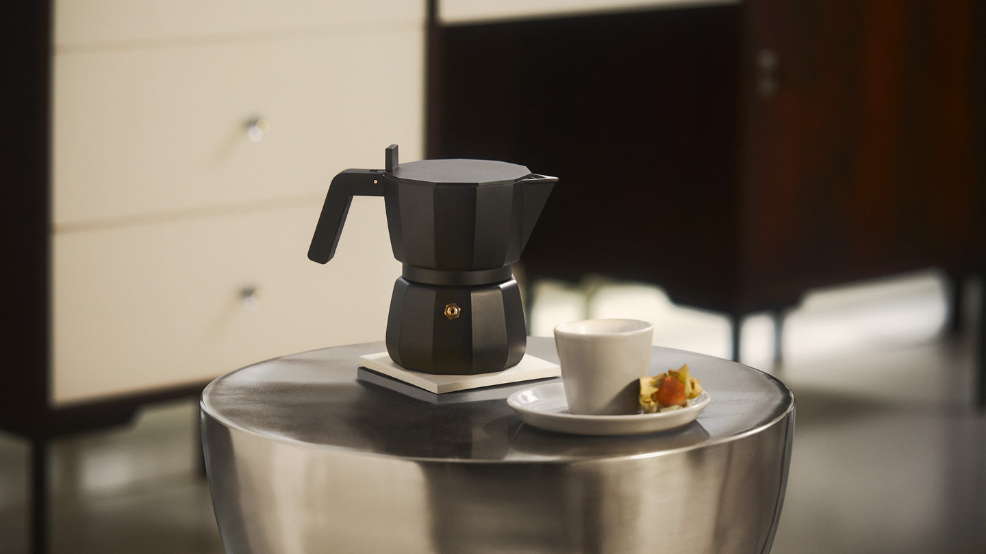 Matte black Moka pot on white coaster beside white cup and saucer with pastry, on metallic table, warm lighting.
