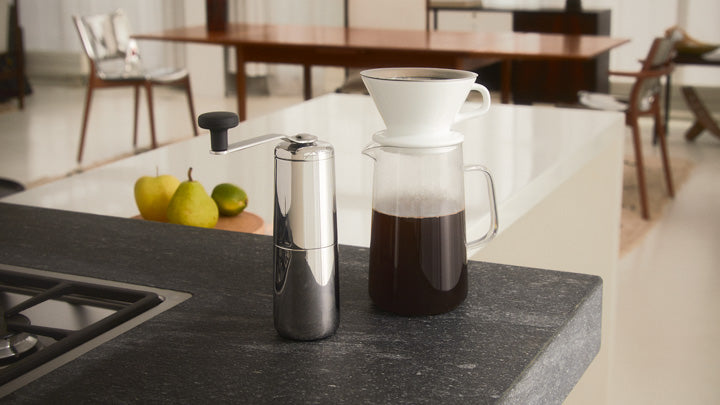 Chrome coffee grinder and glass pour-over brewer on dark countertop, with fruit and dining area in soft-focus background.