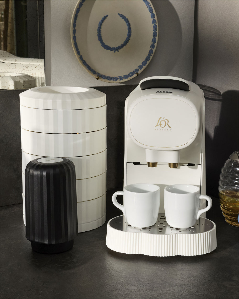 White pleated coffee maker by Alessi, with gold accents, two white mugs on a matching ridged drip tray, and stacked storage containers.