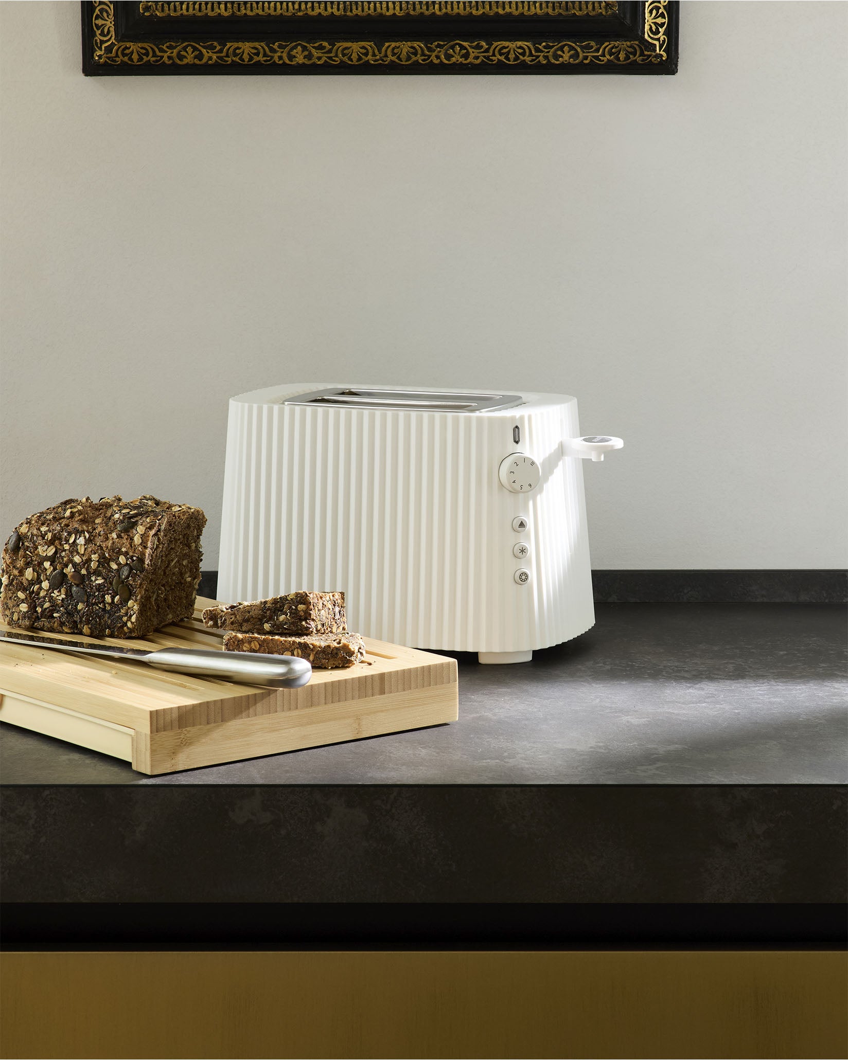 White ribbed toaster by Alessi on a dark countertop with sliced seeded bread on a wooden cutting board.