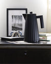 Black electric kettle by Alessi with a distinct pleated texture, styled on a dark dresser with a framed photo and books.
