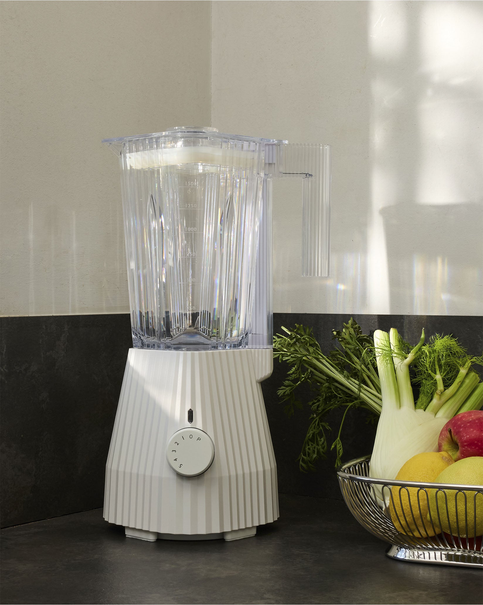 White pleated blender by Alessi with a clear ribbed jar, standing on a dark countertop next to fresh vegetables and fruit in a silver bowl.
