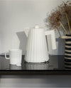 White electric kettle by Alessi, featuring a distinctive fluted Plissé design, styled with a white mug and dried flowers.