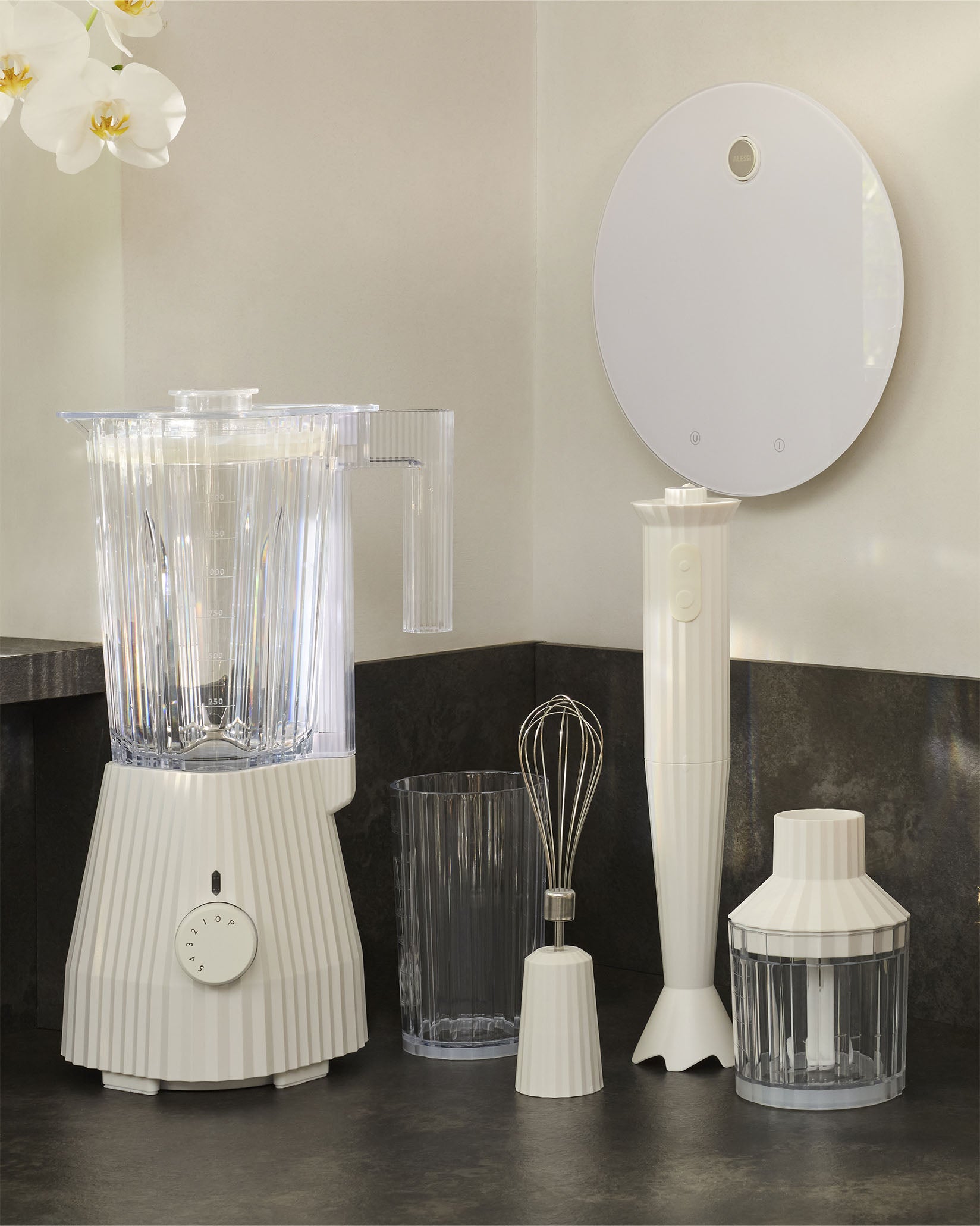 White pleated hand blender by Alessi, part of a matching kitchen appliance collection on a dark countertop.