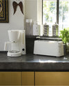 White ribbed toaster by Alessi with a chrome warming rack on a dark countertop in a kitchen.