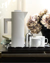 White pleated thermal carafe by Alessi, with handle and spout, shown on a black tray with mugs and dried flowers.