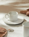 White ceramic espresso cup and saucer by Alessi, with a subtly patterned saucer, on a table with a brown napkin and a silver spoon.