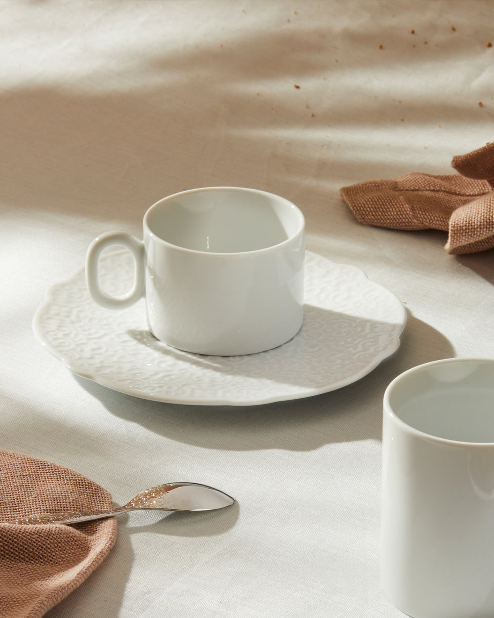 White ceramic espresso cup and saucer by Alessi, with a subtly patterned saucer, on a table with a brown napkin and a silver spoon.