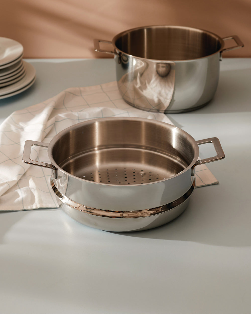 Two shiny stainless steel cooking pots by Alessi, one a steamer pot with a perforated insert, on a blue counter.