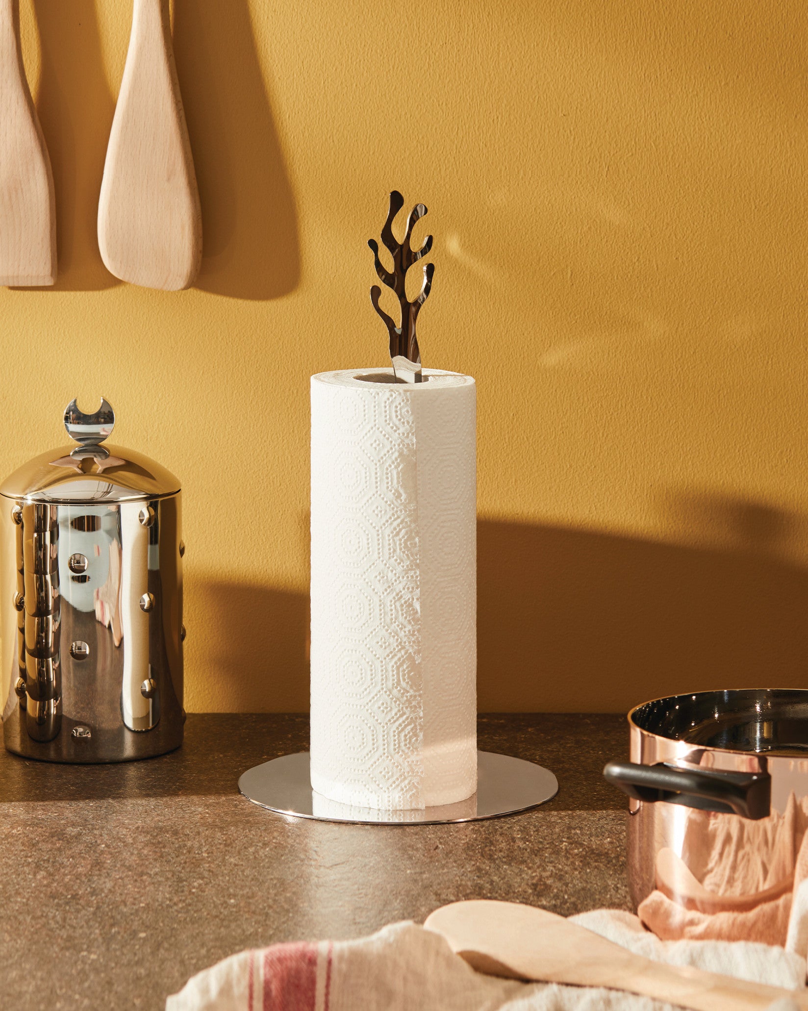 Polished chrome paper towel holder with an abstract coral-shaped top by Alessi, holding a white paper towel roll on a counter.