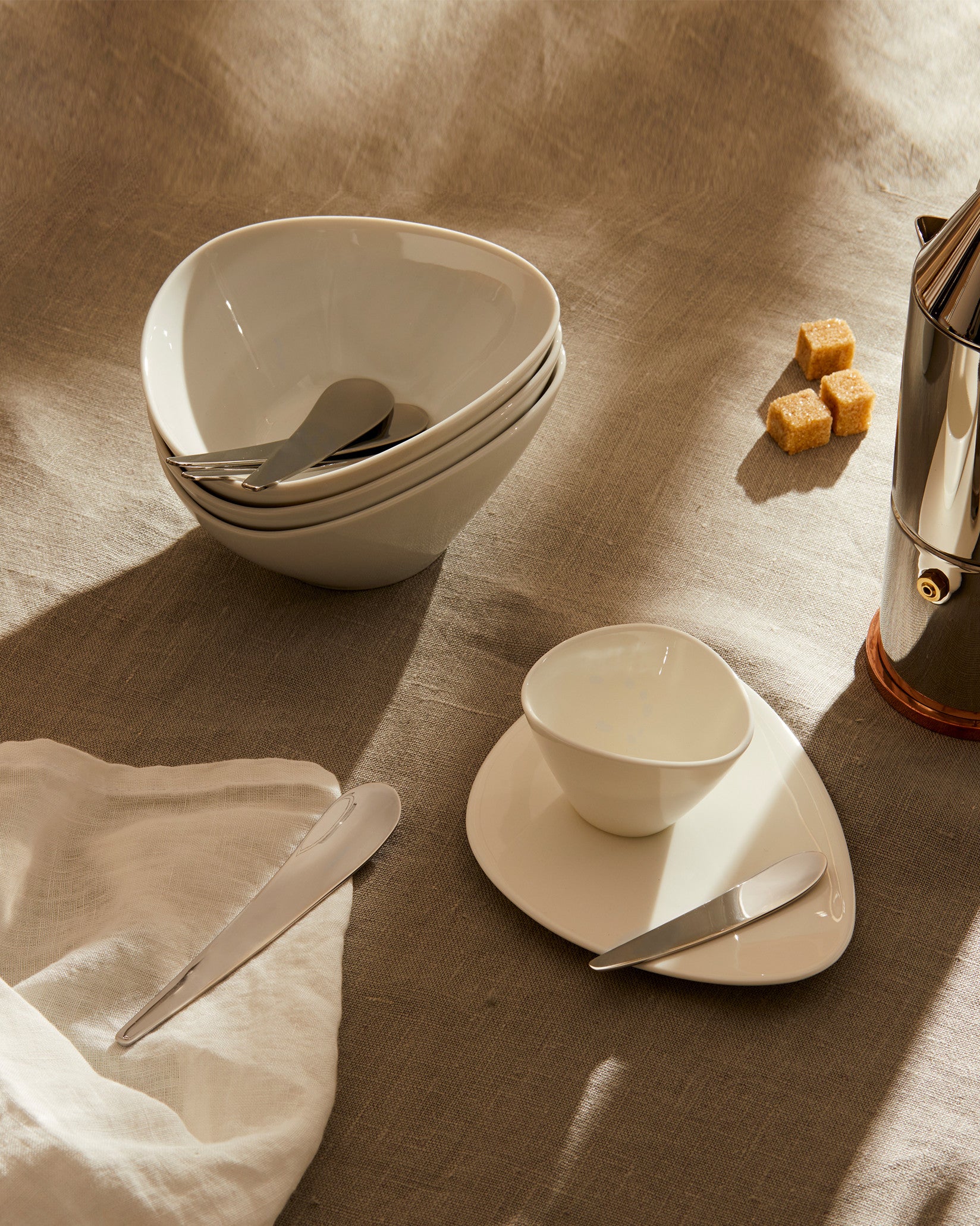 White ceramic Colombina collection by Alessi, featuring irregularly shaped bowls, plates, and modern cutlery on a linen tablecloth.