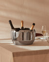 Silver segmented stainless steel champagne cooler by Alessi, holding three bottles on a linen tablecloth.