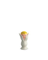 White egg cup by Alessi, featuring hands cradling a pink face with a yellow cap and gold band.