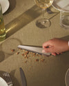 Silver triangular Ala crumb sweeper by Alessi, sweeping food crumbs on a khaki linen tablecloth.