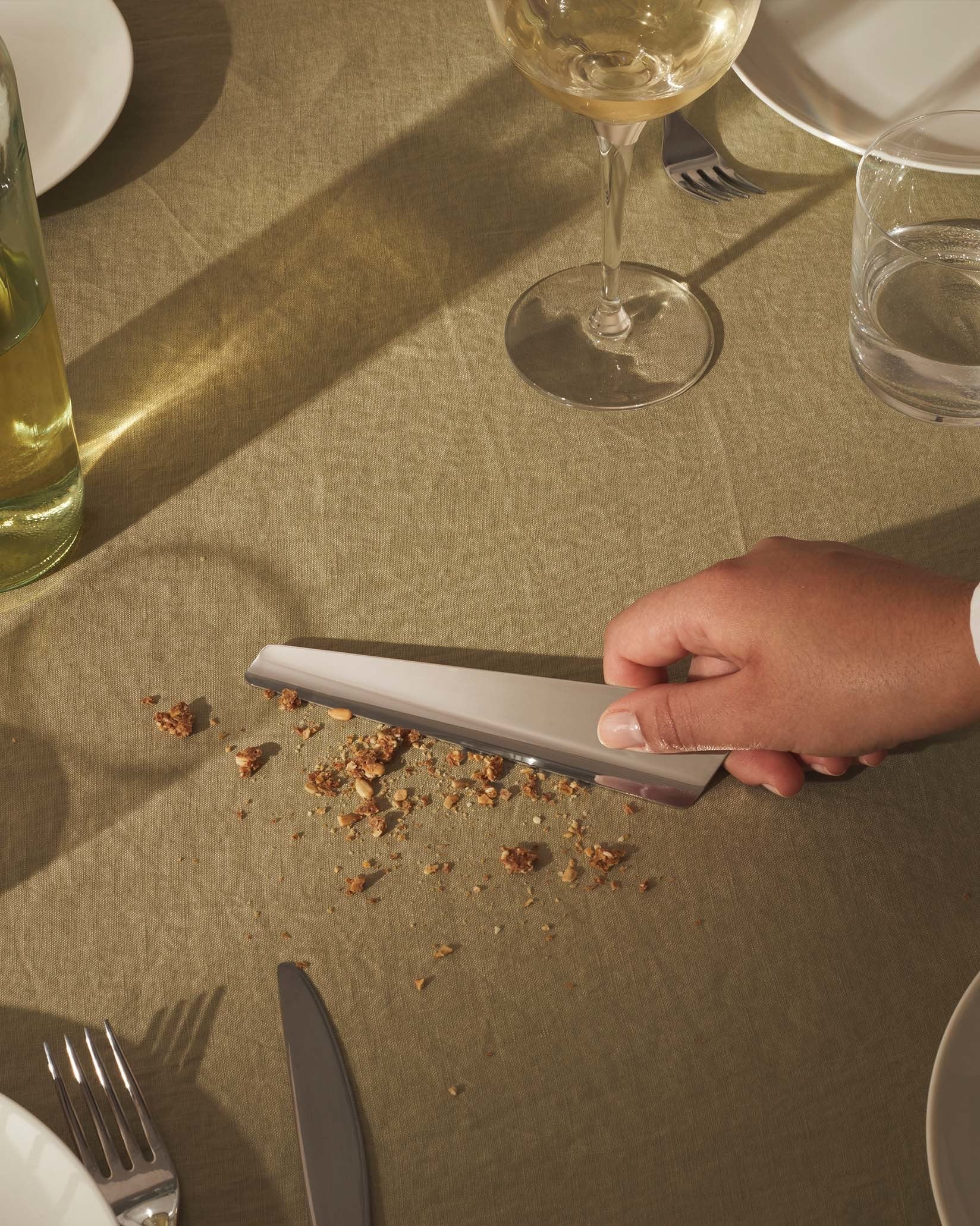 Silver triangular Ala crumb sweeper by Alessi, sweeping food crumbs on a khaki linen tablecloth.