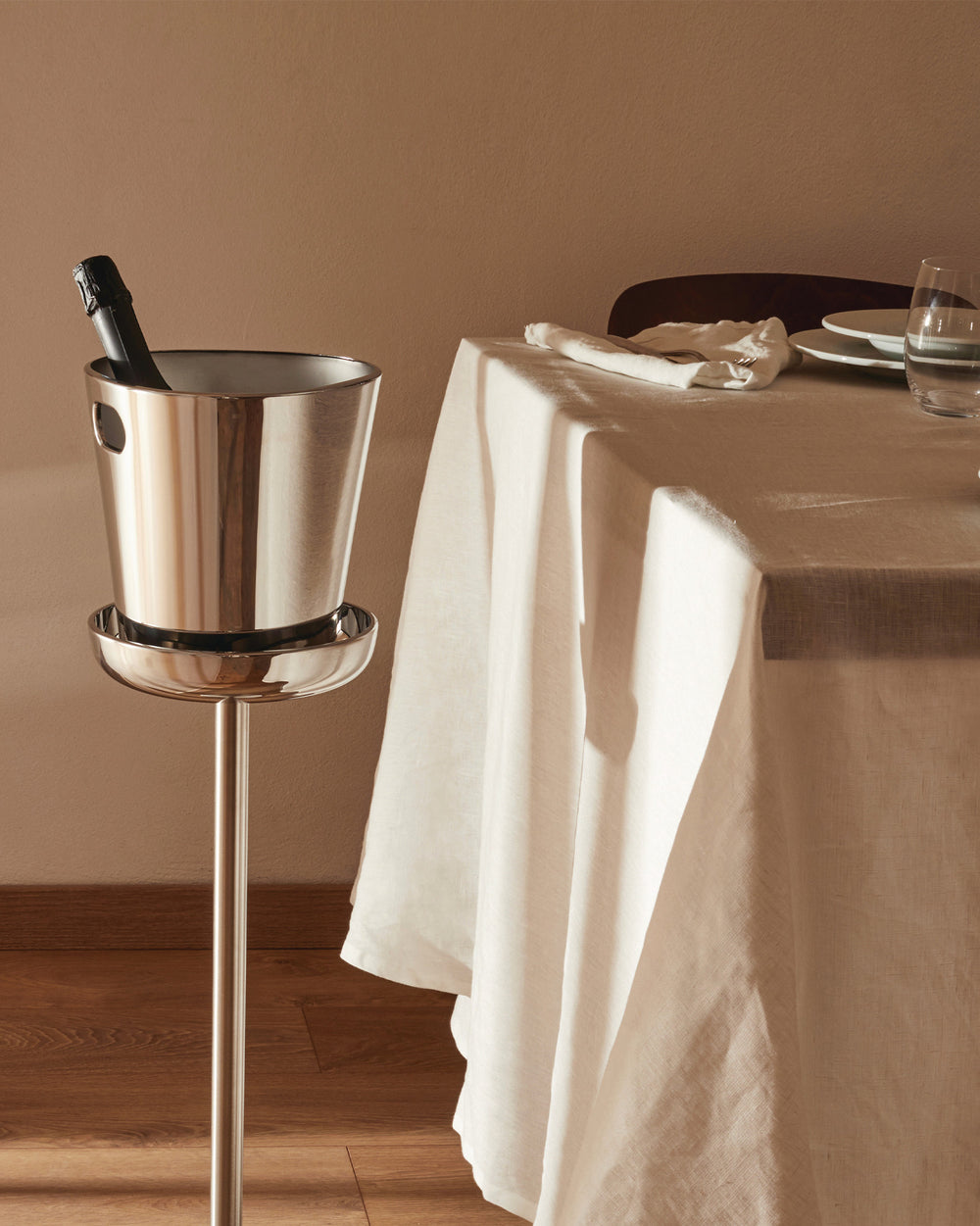 Stainless steel champagne cooler and stand by Alessi, holding a bottle next to a set dining table.