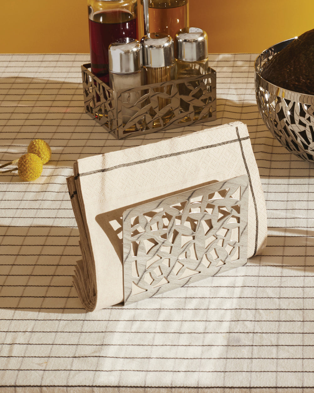 Silver-toned CACTUS! napkin holder by Alessi with abstract cutout design, alongside matching cruet set holder and bowl on a striped tablecloth.