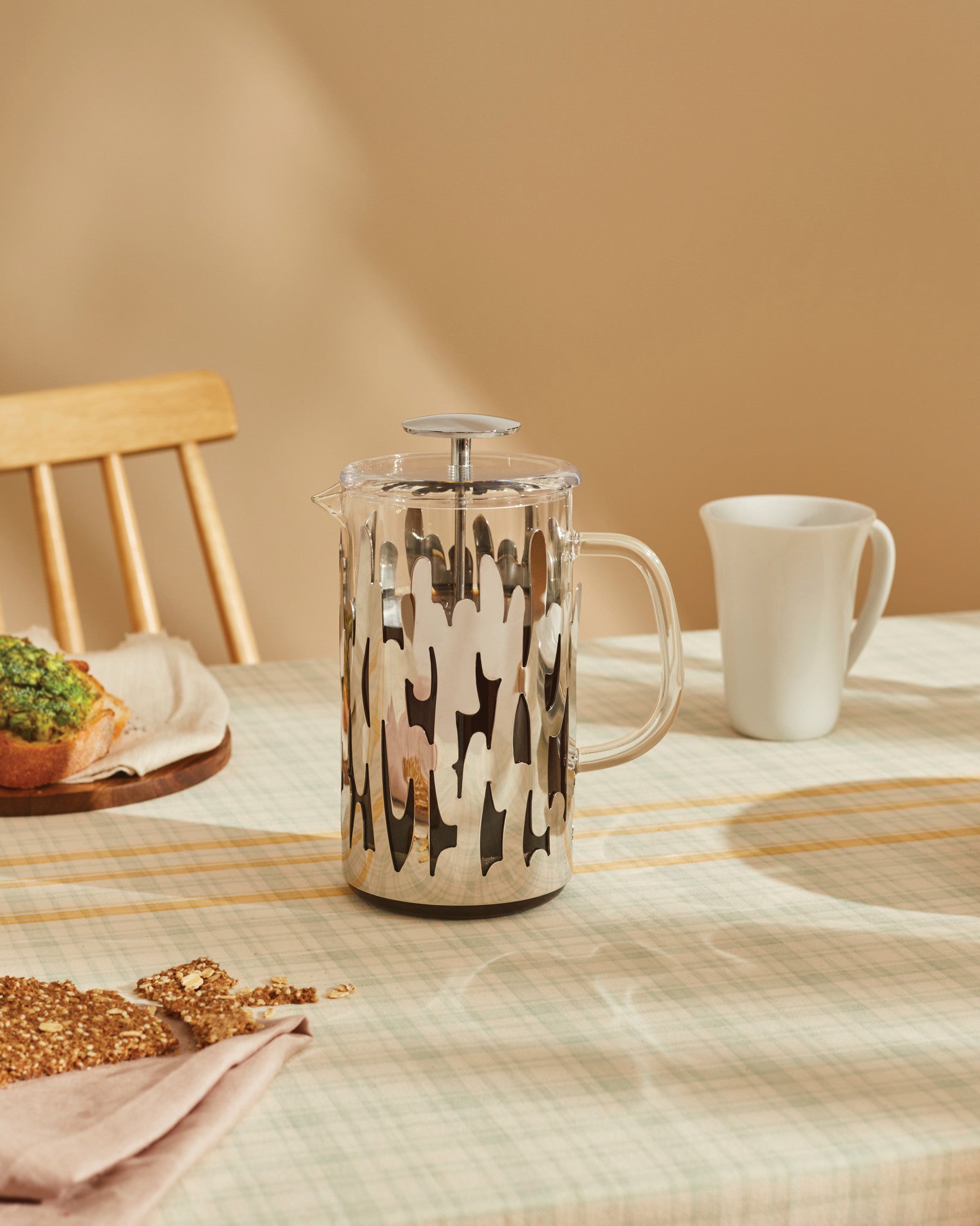 Clear glass French press by Alessi, with a silver metallic bark-like patterned sleeve, on a plaid tablecloth.