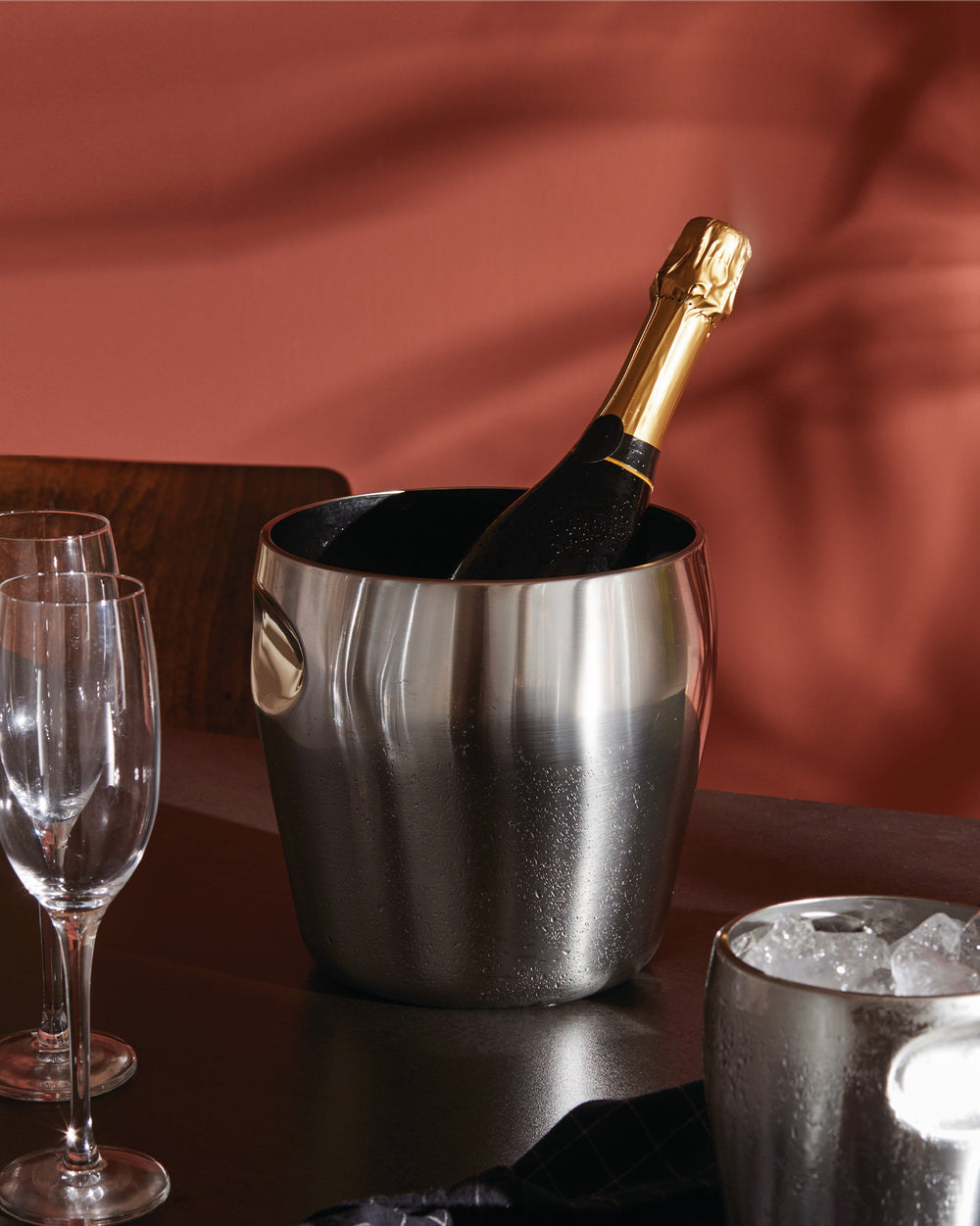 A brushed stainless steel champagne bucket by Alessi, holding a bottle, with an ice bucket and two empty wine glasses.