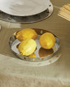 Silver round tray with cut-out figures by Alessi, holding three lemons on a green tablecloth, with plates and spaghetti.