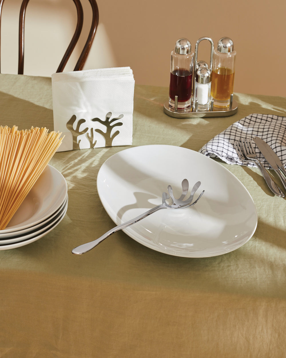 Silver metal spaghetti server with a coral design by Alessi, resting on a white oval plate on a green tablecloth.