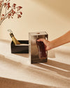 Stainless steel bottle opener and cap collector by Alessi, with a hand placing a dark bottle into its opening.
