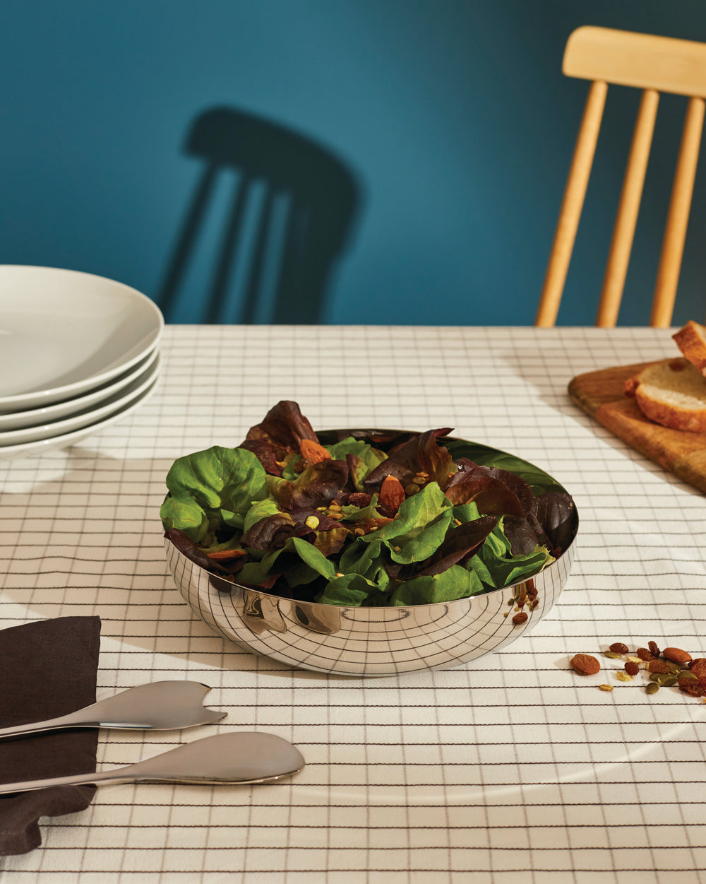 Stainless steel salad bowl by Alessi, filled with fresh mixed greens and nuts, on a grid-patterned tablecloth with serving utensils.