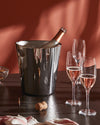 Dark grey metallic ice bucket by Alessi, glistening with water droplets, chilling a rose champagne bottle beside glasses and a cork.