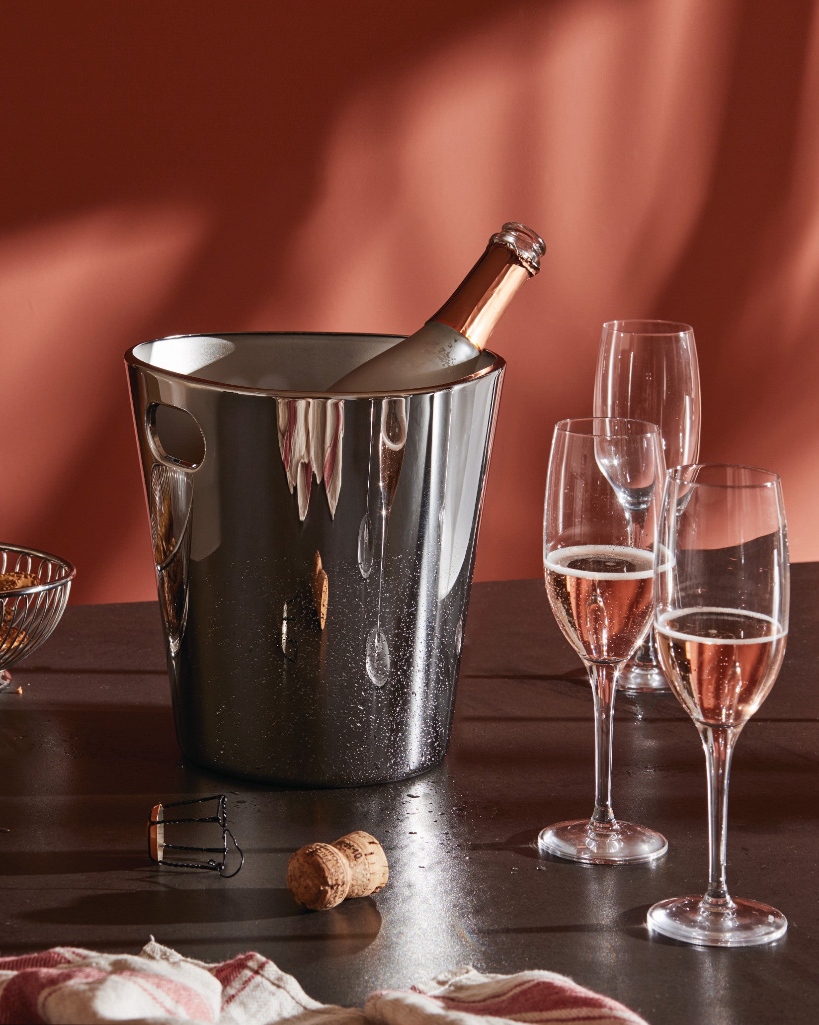 Dark grey metallic ice bucket by Alessi, glistening with water droplets, chilling a rose champagne bottle beside glasses and a cork.