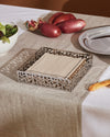 Silver metal napkin holder by Alessi with a geometric cactus cut-out design, holding beige napkins on a linen table runner.