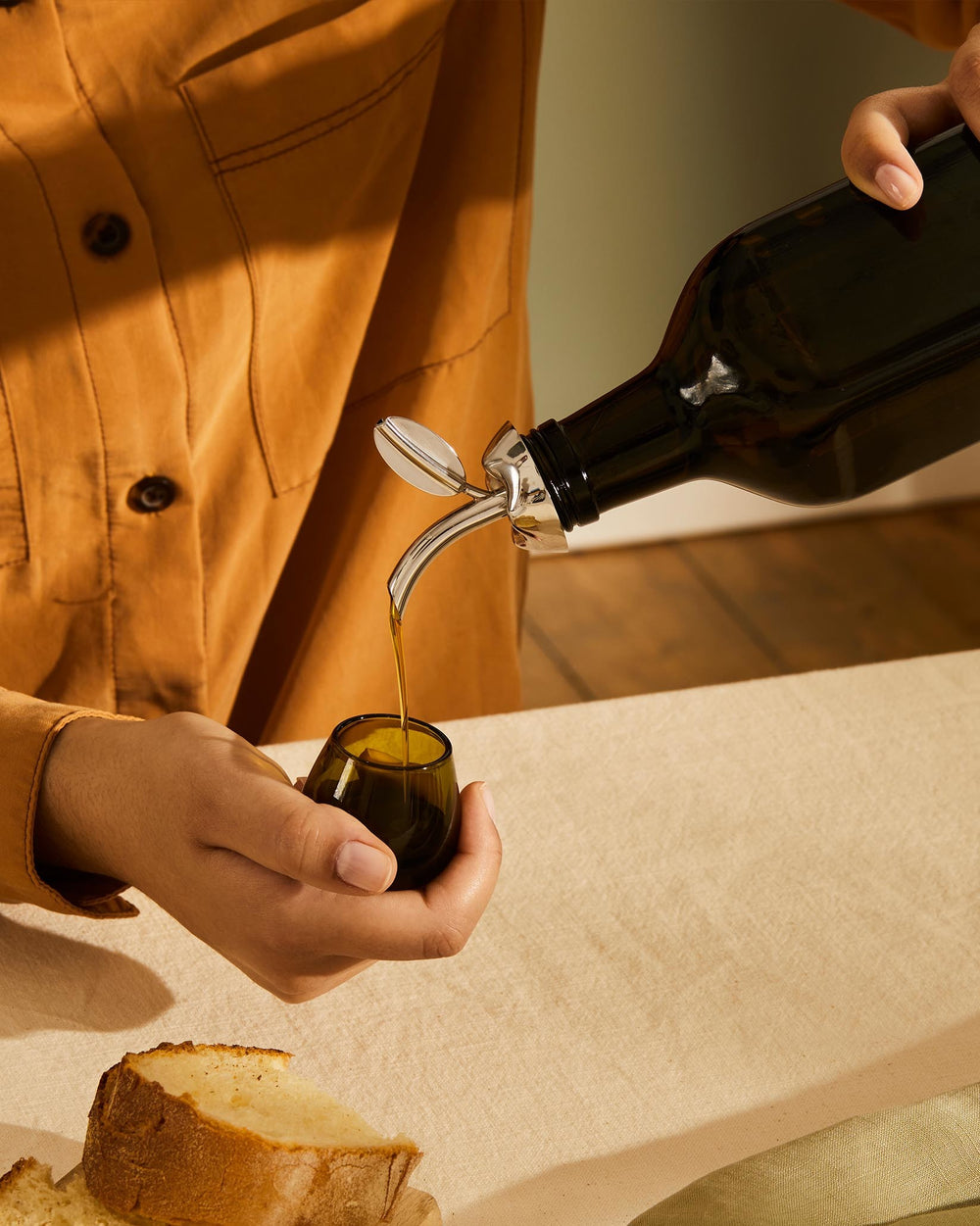 Stainless steel oil pourer by Alessi, dispensing golden olive oil into a small dark glass held by a person.