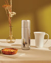 Silver stainless steel vacuum flask by Alessi, upright on a beige tablecloth with dried flowers, a mug, and a tart.