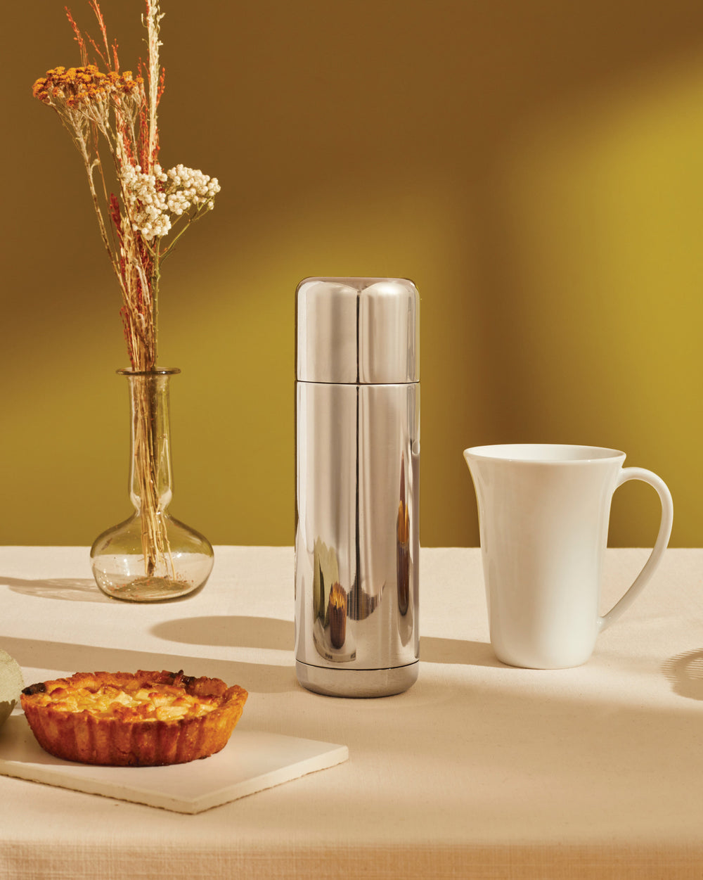 Silver stainless steel vacuum flask by Alessi, upright on a beige tablecloth with dried flowers, a mug, and a tart.