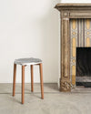 Silver molded seat stool by Alessi with natural wood legs, on a concrete floor beside an antique fireplace.