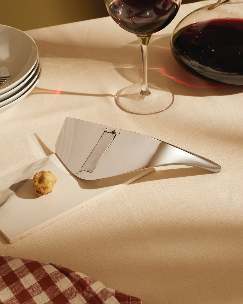 Silver metal truffle slicer by Alessi, resting on a marble board with a truffle, wine glass, and decanter.
