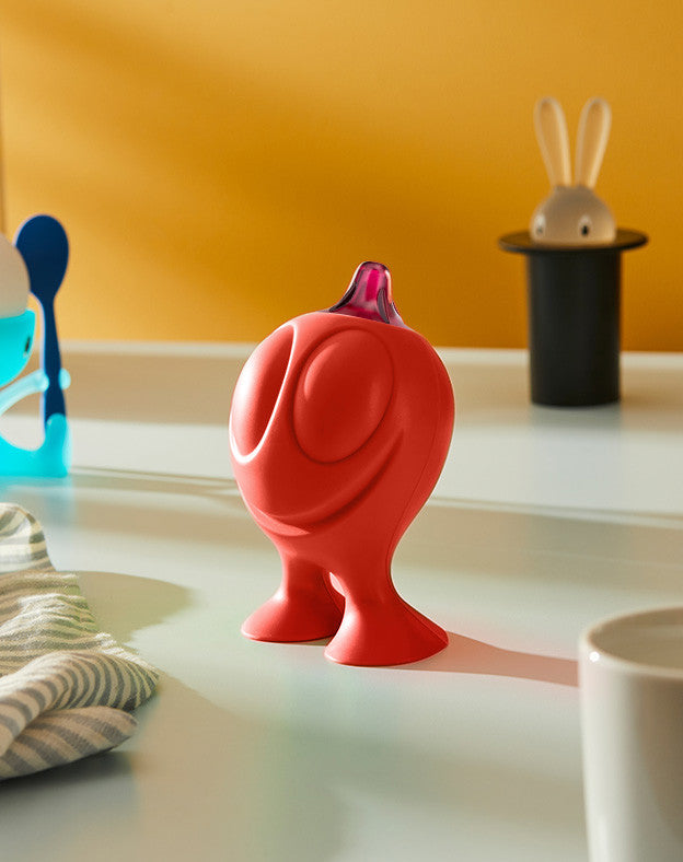 Red Gino Zucchino dispenser by Alessi, designed with a smiling face and purple pour spout, on a counter.