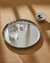 Clear glass pitcher by Alessi filled with bubbly water, on a round silver tray with a glass, beside a modern silver ashtray on a shadowed wood table.
