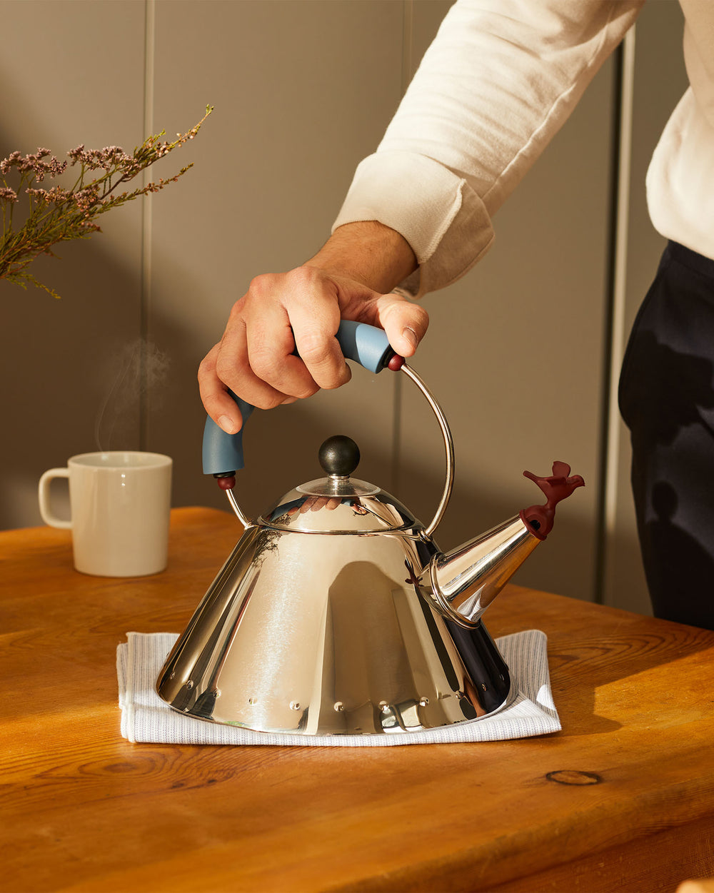 Polished silver kettle by Alessi with a blue handle and red bird whistle, held on a wooden table.