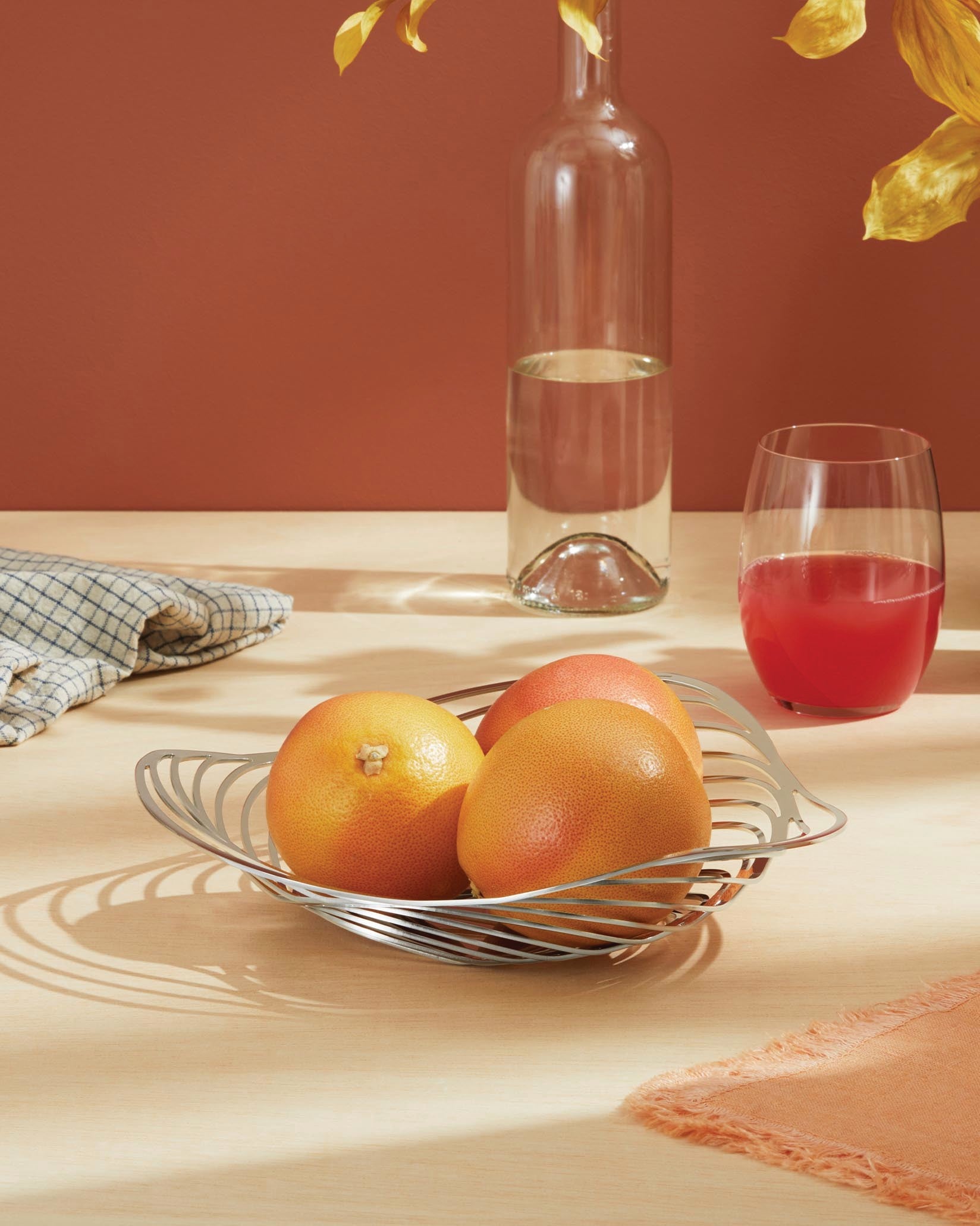 Silver metal fruit bowl by Alessi with a wavy design, holding three grapefruits on a wooden table.
