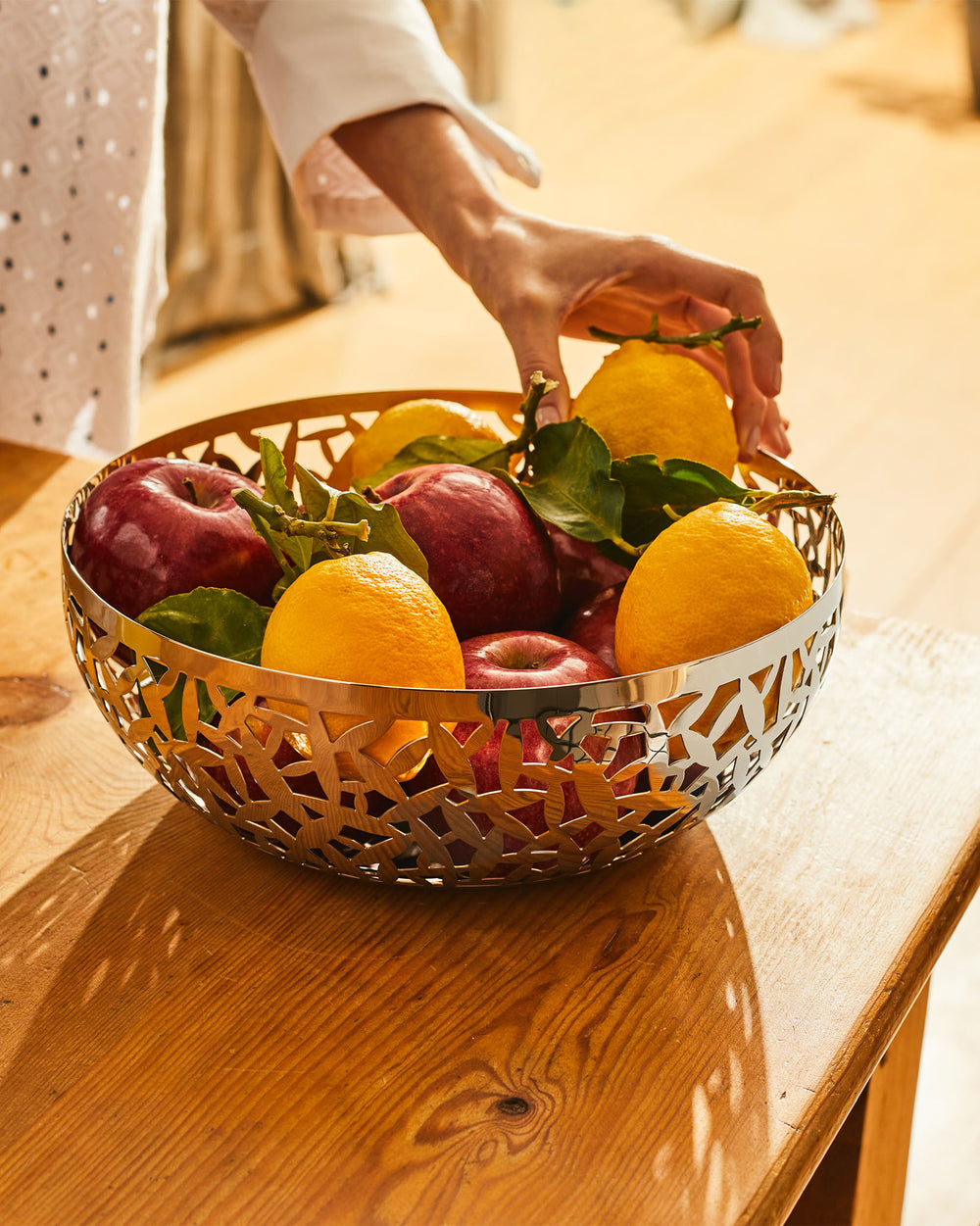 Polished metal fruit bowl by Alessi, featuring an intricate cactus-inspired cut-out design, filled with fresh fruit on a wooden table.