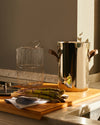 Tall stainless steel pot by Alessi with brown leather handles, alongside a gold wire insert basket and fresh asparagus.