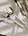 Silver-toned cutlery set by Alessi, featuring a fork and spoon with sleek handles, on a white plate and striped napkin.