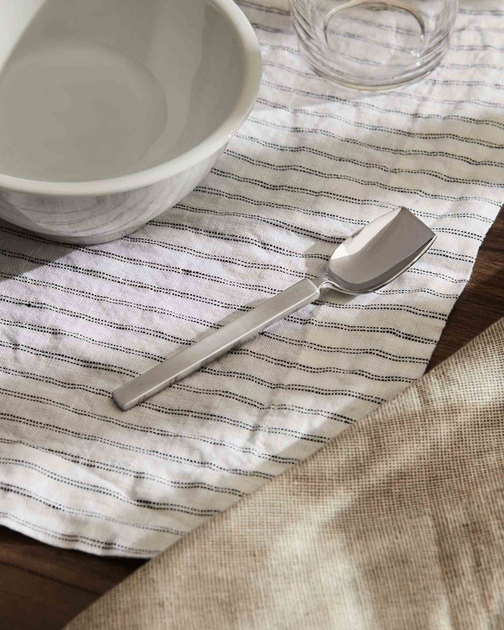 Silver serving spoon by Alessi with a flat handle, on a striped linen cloth with a white bowl and clear glass.