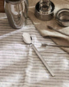 Stainless steel spoon by Alessi with a polished bowl and matte handle, on a striped white linen cloth.