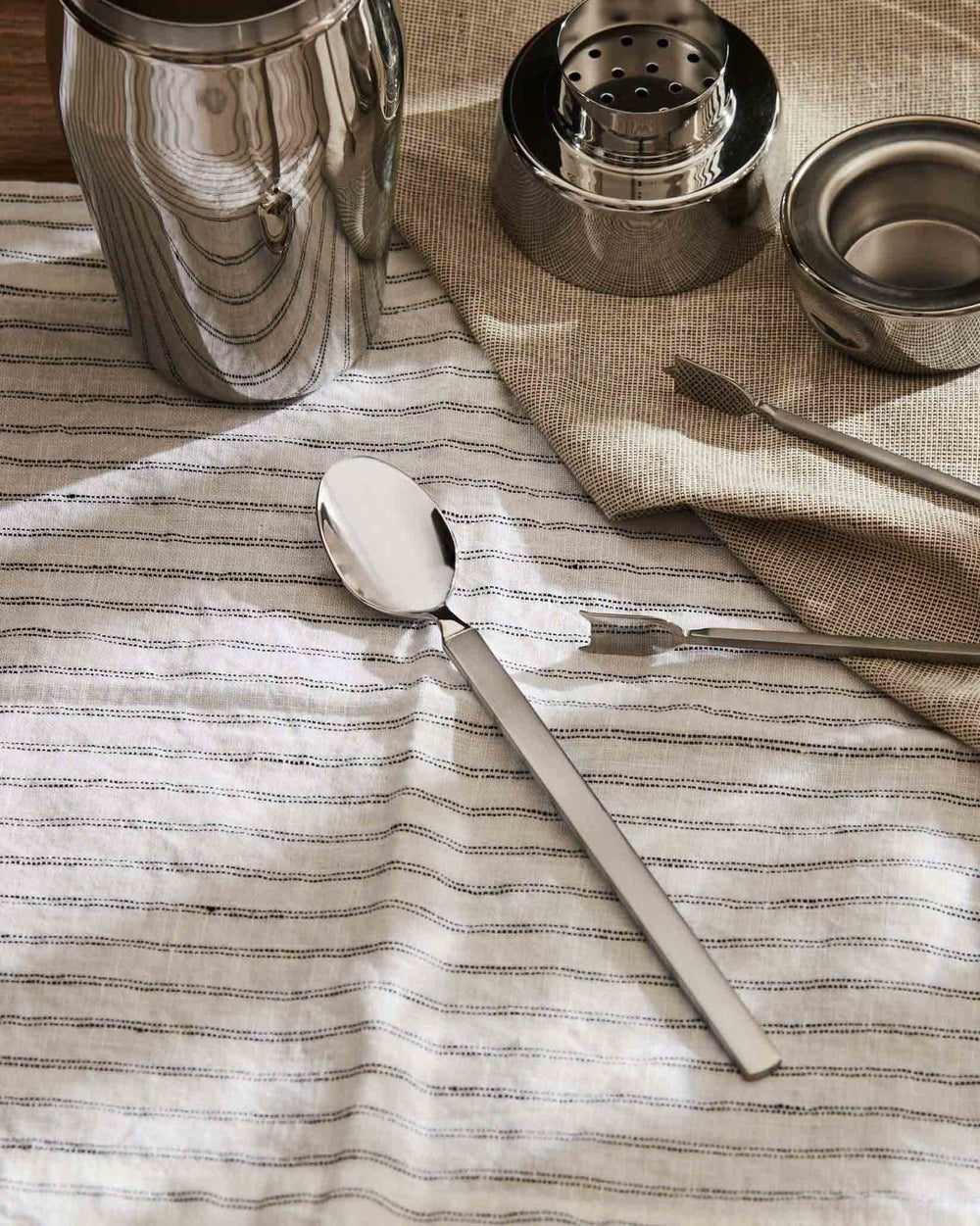 Stainless steel spoon by Alessi with a polished bowl and matte handle, on a striped white linen cloth.